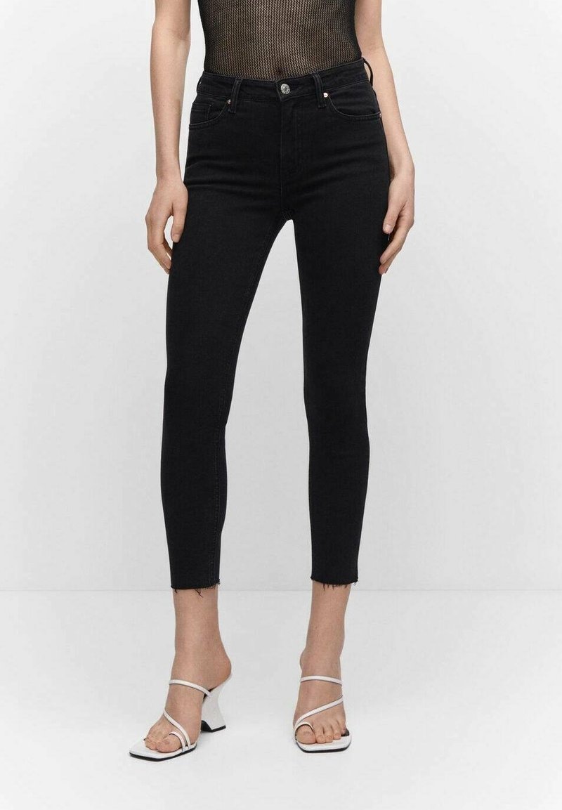 Femme portant un jean skinny noir taille haute avec des ourlets bruts, associé à des sandales blanches à talons et à brides, et un top noir sans manches.
