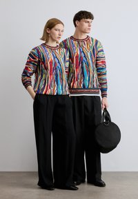 Des pulls en tricot colorés et texturés présentent des motifs tourbillonnants. Le mannequin les associe à un pantalon noir à jambes larges et un sac à main rond noir.