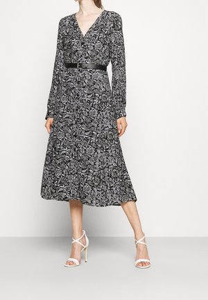 Kobieta ubrana w długą sukienkę midi w czarno-białe kwiaty z czarnym paskiem i białymi sandałami na obcasie, stojąca na jednolitym tle.