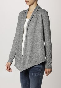 Grå stickad cardigan med öppen framsida, långa ärmar och draperad nederkant. Texturerad tyg med ett subtilt mönster, bärs över en vit topp.