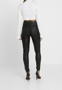 Jeans ajustados de imitación cuero negros con cintura alta, cinco bolsillos y diseño ajustado. Llevados con una blusa blanca de manga larga y tacones altos.
