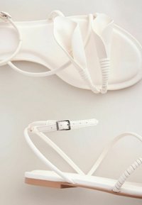 Sandales plates blanches avec fines brides, boucle à la cheville et détail de nœud torsadé sur la bride des orteils, présentées sur un fond neutre.