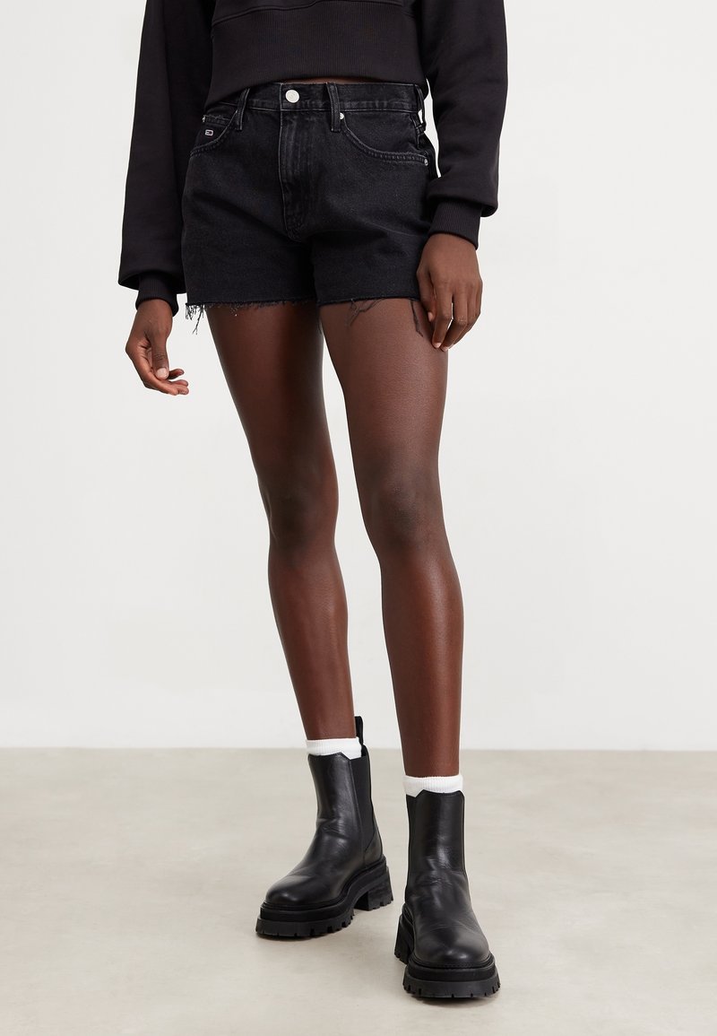 Kvinna klädd i svarta fransiga jeansshorts, svart kort sweatshirt, vita strumpor och svarta klumpiga ankelskor som står på ett ljust golv.