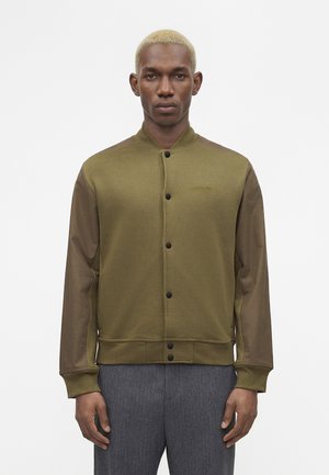 Uomo con capelli corti decolorati che indossa una giacca verde oliva con bottoni e maniche marrone scuro e pantaloni grigi, in piedi davanti a uno sfondo bianco.