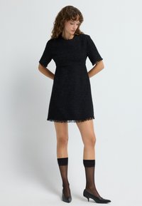 Mustapuku, jossa on teksturoitu, kudottu kangas, lyhyet hihat ja frangipakara. Yhdistetty mustien läpikuultavien polvisukkien ja kärjellä varustettujen korkokenkien kanssa.