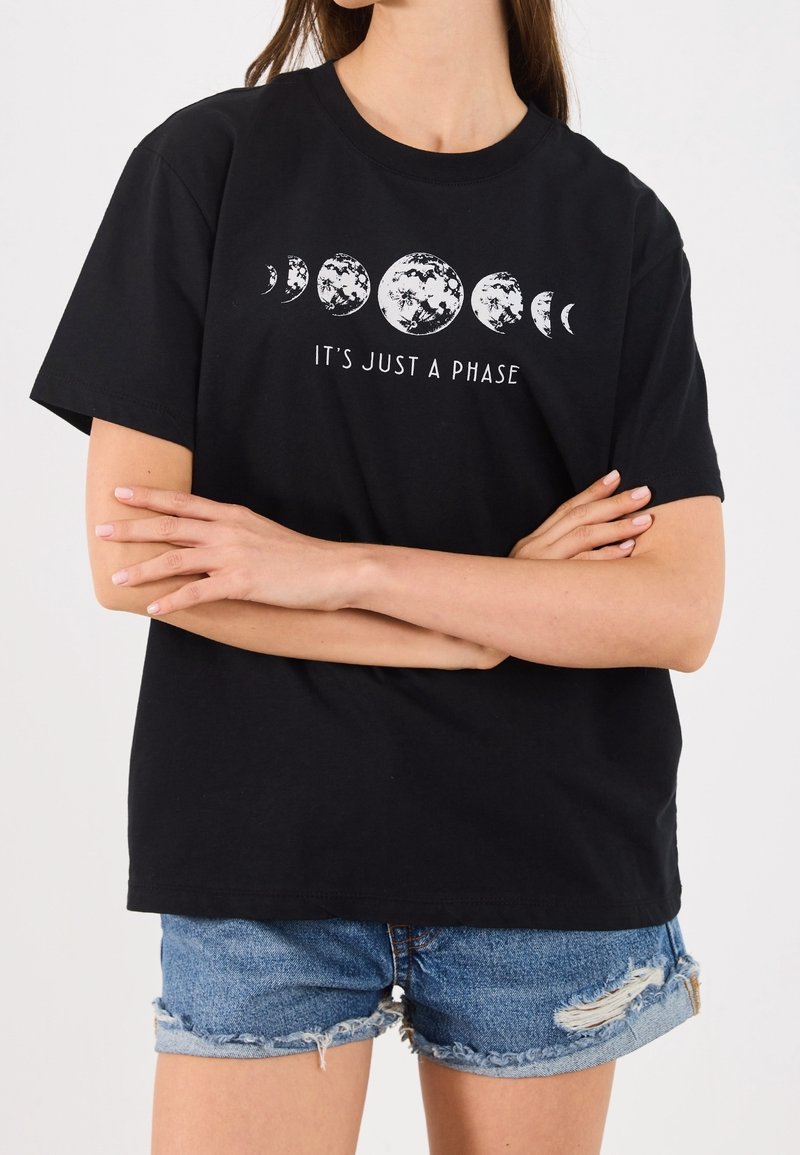 Personne aux bras croisés porte un t-shirt noir montrant les phases de la lune et le texte "It's Just a Phase,", assorti à un short en jean déchiré.