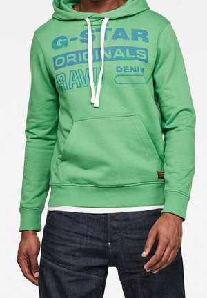 Homme portant un sweat à capuche vert G-Star Originals avec poche avant et cordons blancs, associé à un jean bleu foncé.