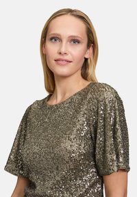 Metaliczna złota bluzka z cekinami, z krótkimi rękawami, okrągłym dekoltem i teksturowaną powierzchnią, która odbija światło, tworząc migoczący efekt.