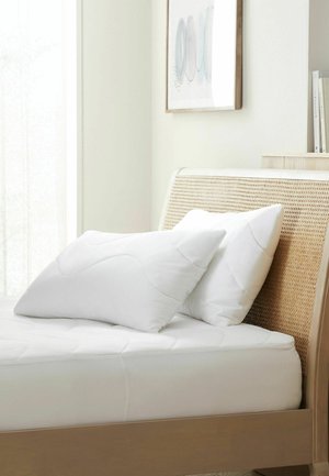Bed met beige geweven hoofdeinde, wit matras en twee witte kussens in een lichte, minimalistische slaapkamer.