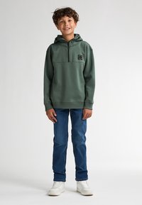 Zelená mikina s kapucí a polozipem, předn�í kapsou a nášivkou s logem, doplněná modrými džíny a bílými teniskami na jednobarevném pozadí.