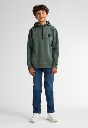 Sweat à capuche vert avec demi-fermeture éclair, poche avant et écusson, associé à un jean bleu et des baskets blanches sur un fond uni.
