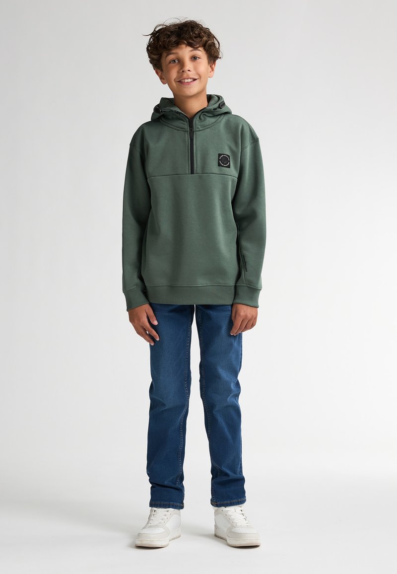 Zelená mikina s kapucí a polozipem, předn�í kapsou a nášivkou s logem, doplněná modrými džíny a bílými teniskami na jednobarevném pozadí.