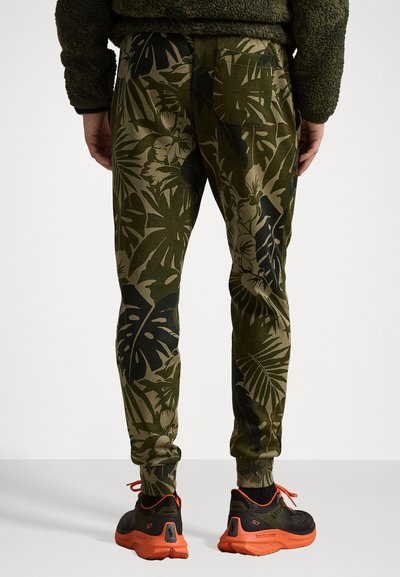 Pantalones de chándal verde oliva con un gran patrón de hojas tropicales verdes oscuras, con tobillos ajustados y un bolsillo trasero, combinados con zapatillas negras y naranjas.