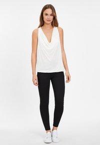 Bílý bezrukávový top s hlubokým výstřihem, kombinovaný s černými těsnými kalhotami a bílými teniskami, na neutrálním pozadí.