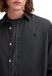 Camisa negra de botones con una textura suave, que presenta una tapeta oculta, botones claros y un pequeño logo bordado en el pecho.