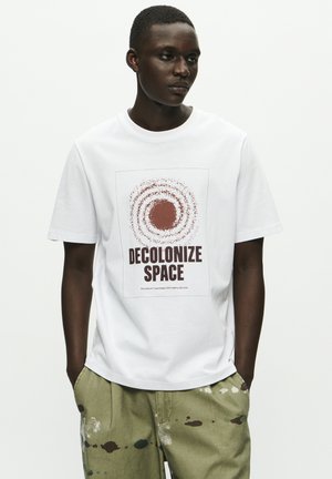 Biała bawełniana koszulka z dużą, okrągłą grafiką w kolorze brązowym z napisem "DECOLONIZE SPACE". Model nosi oliwkowe spodnie z kolorowymi plamkami farby.