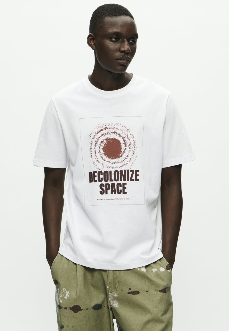 Balta kokvilnas T-kreklu ar lielu, apaļu grafiku brūnā krāsā, kurā ir uzraksts "DECOLONIZE SPACE". Modelis valkā olīvzaļa bikses ar daudzkrāsainiem krāsas pleķiem.