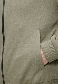 Personne portant une veste olive claire avec fermeture éclair noire, main droite placée dans la poche avant, texture du tissu visible.