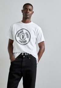 T-shirt blanc à manches courtes avec un graphique circulaire noir portant le texte "VERSACE JEANS COUTURE", associé à un pantalon en denim noir.