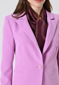 Lila blazer met een gladde textuur, reverskraag en een gouden cirkelvormige knoopaccent. Daaronder is een diep bordeauxrode sjaal gelaagd.