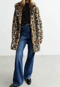 Płaszcz z futra syntetycznego w zwierzęcy deseń lamparta z szerokim kołnierzem, w połączeniu z niebieskimi spodniami typu dzwony o wysokim stanie i błyszczącymi czarnymi butami, na neutralnym tle.