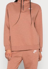 Nike-pullover i dammrosé, med hög krage och halvdragkedja. Mjukt material, löst sittande, med vita logodetaljer på bröstet och midjan.
