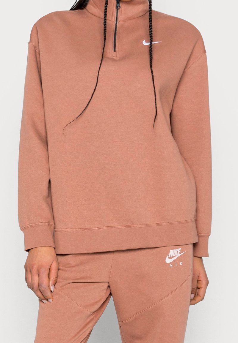 Nike-pullover i dammrosé, med hög krage och halvdragkedja. Mjukt material, löst sittande, med vita logodetaljer på bröstet och midjan.