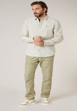 Homme portant une chemise à rayures beige à boutons, un pantalon vert olive clair, et des baskets beige et blanches, debout les mains croisées et regardant vers la droite.