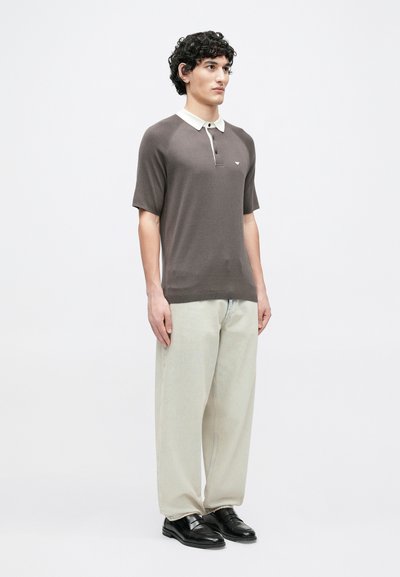 Polo marron tricoté avec col blanc, manches courtes raglan, patte de boutonnage à trois boutons, associé à un pantalon ample de couleur claire et des chaussures noires.