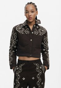 Brązowa krótka kurtka z białym haftem w kwiaty, dwiema przednimi kieszeniami i zapięciem na guzik. W komplecie z pasującymi spodniami, które mają podobne detale.