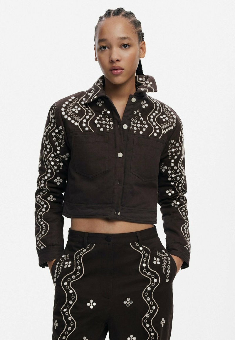 Brązowa krótka kurtka z białym haftem w kwiaty, dwiema przednimi kieszeniami i zapięciem na guzik. W komplecie z pasującymi spodniami, które mają podobne detale.