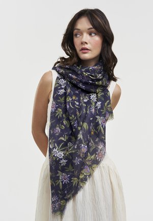 Žena s tmavými vlnitými vlasmi, oblečená v bielych šatách bez rukávov a s veľkou námorníckou šatkou s zelenými a fialovými kvetinovými vzormi.