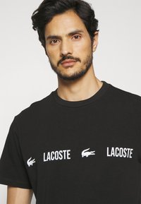 Czarna bawełniana koszulka z krótkim rękawem z trzema białymi logo Lacoste. Posiada okrągły dekolt i luźny krój. Gładka tekstura.