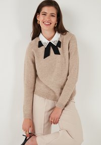 Giovane donna sorridente, con maglione beige con colletto bianco decorato e fiocco nero, pantaloni beige, orecchini dorati e scarpe con tacco nere.