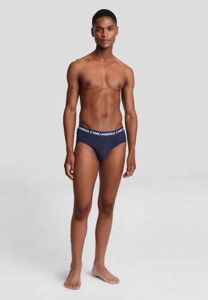 Giovane uomo in piedi a piedi nudi contro uno sfondo semplice, indossa slip blu navy con fascia elastica "KARL LAGERFELD".