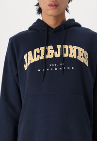 Marineblauwe hoodie van katoen, met een voorste kangoeroezak, een capuchon met trekkoorden, en een opvallende gele logo-opdruk met de tekst "JACK & JONES" boven de borst.