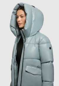 Chaqueta acolchada de material sintético azul claro, con una gran capucha, cierre mediante cremallera y un bolsillo lateral con un parche de logo.