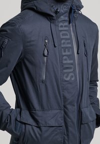 Granatowa kurtka z kapturem, cztery kieszenie na zamek, kieszenie na klapki oraz napis "SUPERDRY" wydrukowany pionowo wzdłuż zamka z przodu.