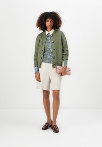 Cardigan verde a maglia con grandi bottoni, top stampato blu, pantaloncini crema e pochette rosa. Mocassini marroni con dettagli in hardware dorato.