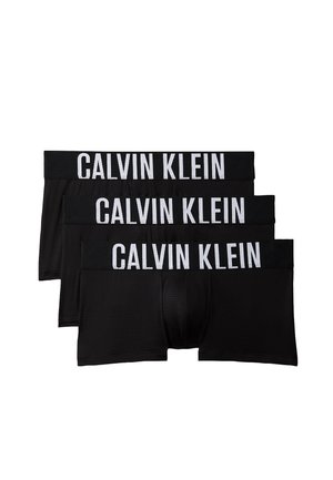 Hemdloser Mann steht barfuß vor einfachem Hintergrund, trägt schwarze Calvin Klein Boxershorts und hält den Bund mit einer Hand.