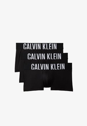Mężczyzna bez koszulki stojący boso na jednolitym tle, ubrany w czarne bokserki Calvin Klein, trzymający pas jedną ręką.