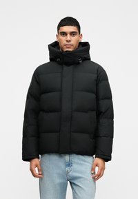 Juodas pūkinis švarkas su gobtuvu, turintis siuvinėtas paneles ir priekį užtrauktą užtrauktuku. Dėvimas virš šviesiai mėlynų džinsų, demonstruojantis laisvą siluetą.