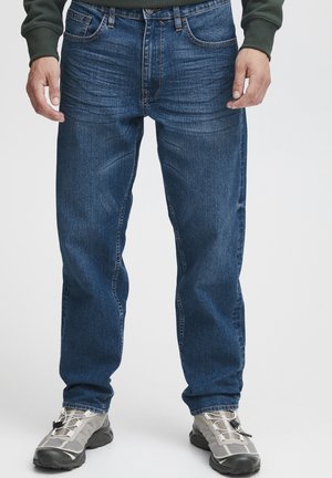 Jeans de mezclilla azul con un corte recto, leve desgaste en los muslos, diseño de cinco bolsillos y herrajes de latón. Llevados con zapatos deportivos grises.