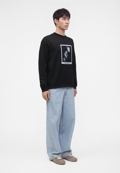 Homme debout portant un sweat-shirt noir avec une impression de portrait en noir et blanc, un jean large bleu clair et des chaussures marron décontractées.