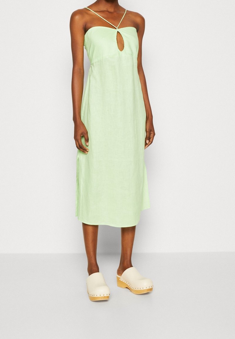 Hellgrünes Leinenkleid mit Schlüsselloch-Ausschnitt und dünnen Trägern, kombiniert mit beigen Holzclogs. Schlichte, lockere Form mit Seitenschlitzen.