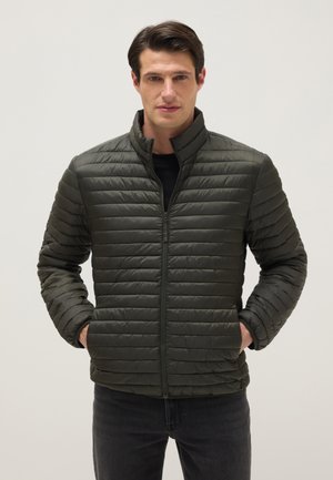 Man draagt een donkergroene doorgestikte jas met ritssluiting, staat met handen in de zakken en een neutrale gezichtsuitdrukking tegen een effen achtergrond.