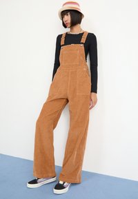 Brown corduroy overalls with wide legs, adjustable shoulder straps, and front pockets. Paired with black and purple sneakers.