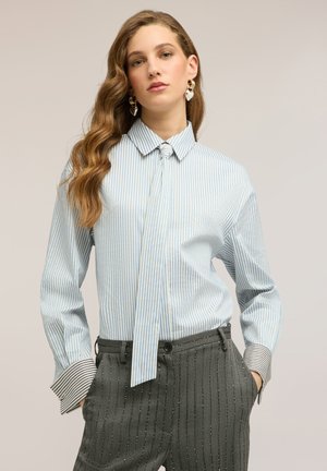 Frau mit welligem Haar trägt eine blau-weiß gestreifte Bluse mit Schleifenkragen und kontrastierenden gestreiften Manschetten, kombiniert mit hoch taillierten grauen Nadelstreifenhosen.