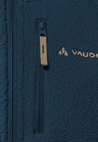 Tumesinine fliiskampsun tekstuurse kangaga, millel on tõmblukuga tasku ja beezist viimistlusest detailid. Brändi logo on esitatud kuldse värviga.