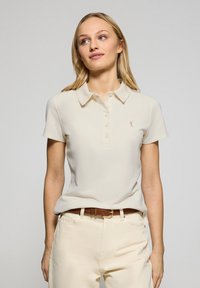 Polo shirt beige con colletto e maniche corte, caratterizzato da una fila di bottoni e un piccolo logo sul lato sinistro del petto. Indossato con pantaloni di colore chiaro.